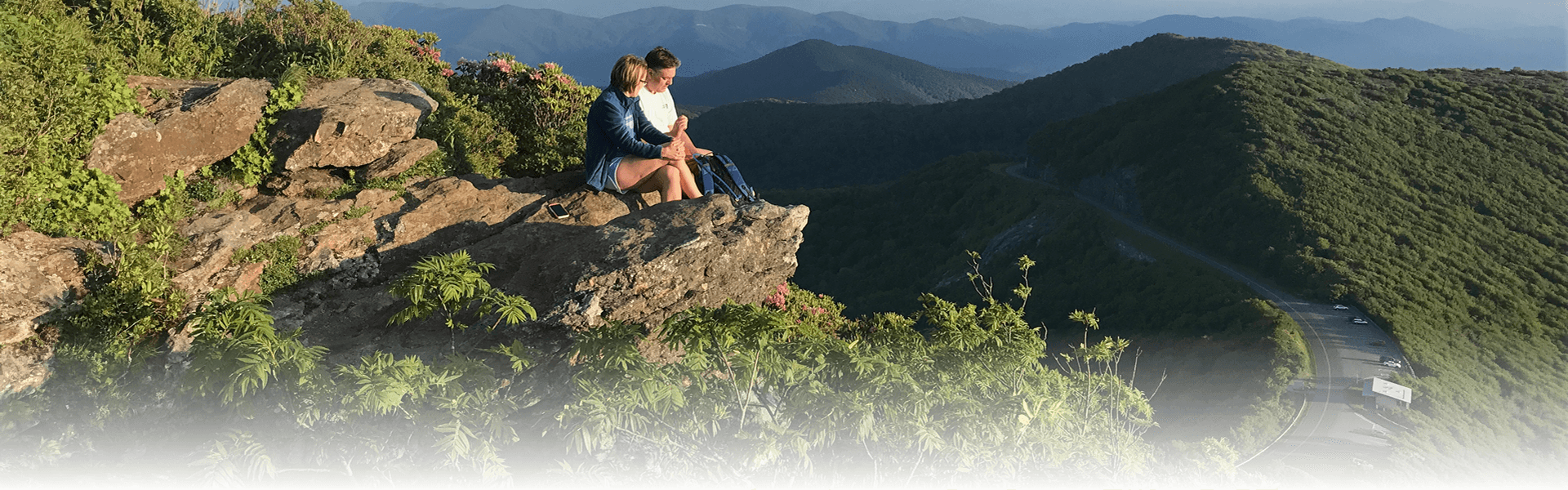 A man and woman sitting at the top of a mountain peak with a view of the mountains