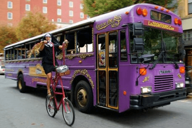 Large Purple Tour Bus with Man on Tall Bicycle dressed as a nun riding beside